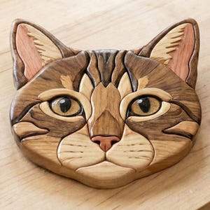 May include: Wooden cat face sculpture with detailed grain patterns. The cat's face is crafted from various wood tones, including light tan, brown, and reddish-brown. The eyes are large and expressive, with a light brown iris and dark pupils. The nose is a soft pink.