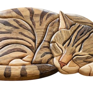 May include: Wooden cat sculpture in a sleeping pose. The cat is carved from various shades of wood, with darker brown stripes on a lighter brown body. The cat's eyes are closed, and its paws are tucked in. The sculpture is a decorative item.