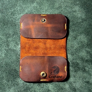 Könnte beinhalten: Offener Kartenhalter aus braunem Leder mit Druckknopfverschluss und gelben Nähten. Der Kartenhalter trägt den Markennamen "Craftano" und ein Bärenlogo.
