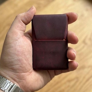 Könnte beinhalten: Ein weinroter Kartenhalter aus Leder mit Klappe. Die kleine Geldbörse wird in einer Hand gehalten und zeigt die satte Farbe und Textur des Materials. Das Design ist minimalistisch und kompakt, geeignet für wichtige Karten.