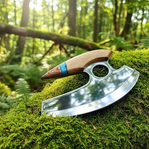 May include: A handcrafted, curved blade knife with a wooden handle and a turquoise inlay. The polished steel blade has a unique shape with a finger hole. The knife rests on a bed of green moss, with a forest background.