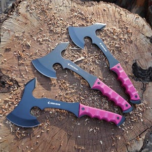 May include: Three black hatchets with pink handles are arranged on a wooden surface. The hatchets have a curved blade and a textured handle. The text "Deer Edge" is printed on the hatchet.