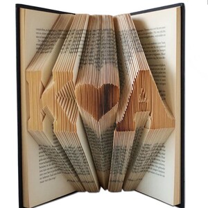 May include: A hardback book with pages folded to spell "I ❤️ U" and a heart shape. The book's pages are beige, and the cover is black. The folded pages create a 3D effect, showcasing the message.