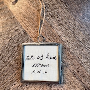 Könnte beinhalten: Ein quadratischer Hängerahmen mit silberfarbenem Metallrand und Glasfront. Im Inneren befindet sich ein weißer Stoffeinsatz mit schwarzer, gestickter Schreibschrift, die "lots of love mum xxx" lautet. Eine braune Kordelschlaufe ist oben befestigt.