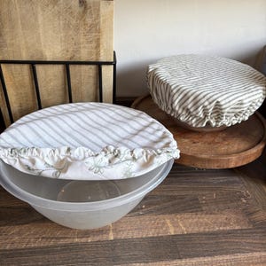 May include: Two round fabric bowl covers with elasticated edges. One cover features a white and grey striped pattern, while the other displays a white and grey striped pattern combined with a light green botanical print of leaves and flowers.