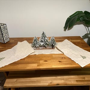 Puede incluir: Una mesa de madera con un camino de mesa claro y árboles de Navidad decorativos. Dos caminos de mesa a juego con diseños de árboles bordados a cada lado. Una lámpara decorativa con un diseño floral está a la izquierda.