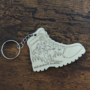 Puede incluir: Llavero de madera con forma de bota de senderismo. La bota presenta un diseño grabado de un excursionista, árboles y montañas, con las palabras "Adventure awaits...". Incluye una cadena y un anillo plateados.