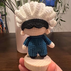 Puede incluir: Muñeca de ganchillo hecha a mano con un tocado blanco con volantes, una cubierta para los ojos negra y un mono azul. La muñeca está montada sobre una pequeña base de madera redonda. Los brazos y la cara de la muñeca son beige.