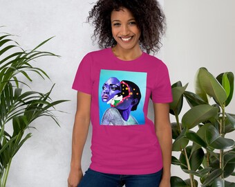Puede incluir: Una camiseta magenta con una vibrante impresión artística de dos caras, una morada y la otra con un patrón negro y dorado. La obra de arte está sobre un fondo azul claro. La modelo lleva vaqueros azules.