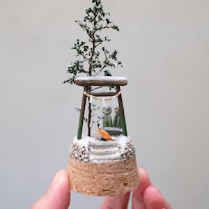 May include: Miniature Japanese torii gate scene with a snow-covered path, two stone statues, and a small orange fox. A snow-covered tree sits atop the gate. The base is a cork cylinder.