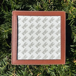 May include: Square brown leather coaster with stitched edges. The center features a white surface with the repeated text "3M" in gray. The coaster is set against a backdrop of green foliage.