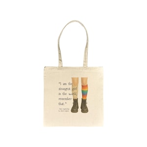 May include: Natural canvas tote bag featuring a quote and illustration. The quote reads "I am the strongest girl in the world, remember that." The illustration shows legs wearing boots and a rainbow-striped sock. The bag has two handles.