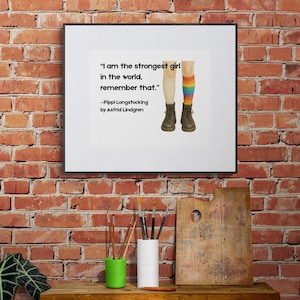 May include: A framed print featuring the quote "I am the strongest girl in the world, remember that." by Astrid Lindgren. The image shows two legs, one with a beige sock and the other with a rainbow striped sock, wearing brown boots. The print is on a wooden table.
