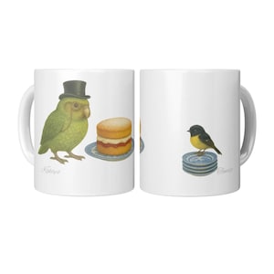 Könnte beinhalten: Weiße Keramik-Tasse mit Illustrationen von Vögeln und Kuchen. Eine Seite zeigt einen grünen Papagei mit Zylinder und Kuchen auf einem Teller. Die andere Seite zeigt einen kleinen Vogel auf einem Stapel Teller. Die Worte "Kakapo" und "Tomer" sind auf der Tasse geschrieben.