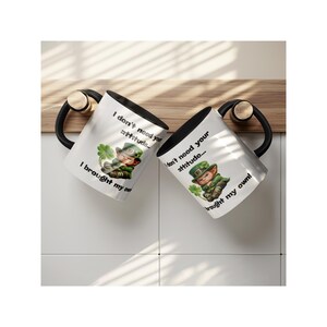 Op de afbeelding: Twee witte keramische mokken met zwarte binnenkant en handvatten. Elke mok heeft een afbeelding van een kabouter met een klavertjevier en de tekst "I don't need your attitude... I brought my own!"