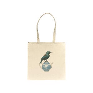 Puede incluir: Bolso tote color crema con un pájaro turquesa posado sobre una tetera azul y blanca. El pájaro, con pico oscuro, mira hacia la izquierda. La tetera tiene un diseño detallado. El bolso tiene dos asas beige.