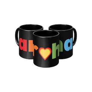 Può includere: Tre tazze in ceramica nera con la parola "aroha" in lettere colorate. La tazza centrale presenta un motivo a cuore. Le tazze hanno una finitura lucida e un manico curvo.