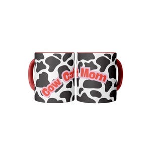 Op de afbeelding: Twee witte keramische mokken met een zwart koeienpatroon en rode handvatten. De tekst "Cow Cat Mom" is in roze op de voorkant van elke mok gedrukt. De mokken zijn ontworpen voor warme of koude dranken.