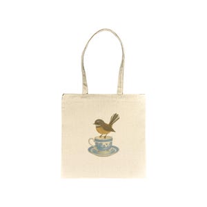 Puede incluir: Bolso tote de color crema con un estampado de un pájaro marrón posado sobre una taza y un platillo azules y blancos. El bolso tiene dos asas y está hecho de lona. El diseño es una ilustración.