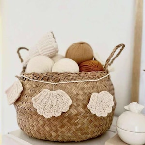 May include: A woven basket filled with yarn balls in shades of white, tan, and brown. The basket is decorated with crocheted seashell ornaments. The basket has two handles and is sitting on a white surface.