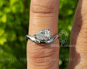 May include: A silver ring featuring a pear-cut diamond. The ring's band has a flowing design, supporting the diamond. The ring is shown on a finger, with a blurred green backdrop. The ring is from BINILA Jewels.
