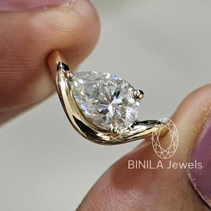 May include: A gold ring featuring a pear-shaped diamond. The ring's band curves around the diamond, creating a unique and elegant design. The ring is held between fingers, showcasing its sparkle. The brand name "BINILA Jewels" is visible.