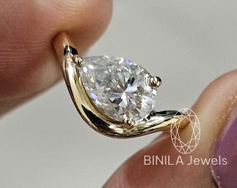 May include: A gold ring featuring a pear-shaped diamond. The ring's band curves around the diamond, creating a unique and elegant design. The ring is held between fingers, showcasing its sparkle. The brand name "BINILA Jewels" is visible.