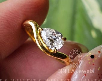 May include: A gold ring featuring a pear-cut diamond. The ring has a distinctive, curved design. The diamond is clear and brilliant. The ring is set against a backdrop of green foliage.