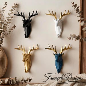 Könnte beinhalten: Vier dekorative Hirschkopf-Wandhalterungen in Schwarz, Weiß, Gold und Blau. Jeder Kopf hat goldene Geweihe und einen Haken unter dem Hals. Die Köpfe sind an einer neutralen Wand befestigt. Unten rechts steht der Text "Faune And Design".