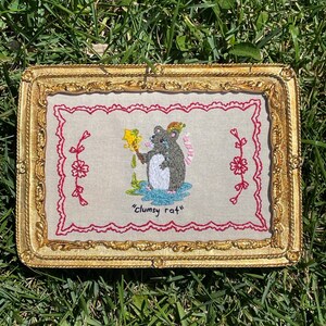 May include: Embroidered artwork in a gold frame featuring a gray rat with a colorful hat, holding a yellow object, and crying. The artwork is framed with a red scalloped border and the text "clumsy rat".