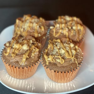 May include: Four chocolate cupcakes in light brown paper liners arranged on a white plate. Each cupcake is topped with a swirl of chocolate frosting, a drizzle of peanut butter, and a sprinkling of crushed peanuts or cookie crumbs on top.
