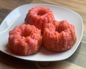 May include: Three pink mini bundt cakes on a white plate. The cakes have a light pink color with a slightly textured surface and a hole in the center. The plate is on a wooden surface.