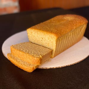 May include: A loaf of golden-brown bread, partially sliced, rests on a white cardboard plate. The bread has a textured crust and a soft interior, suggesting a homemade or artisan quality. The image is well-lit, highlighting the bread's details.