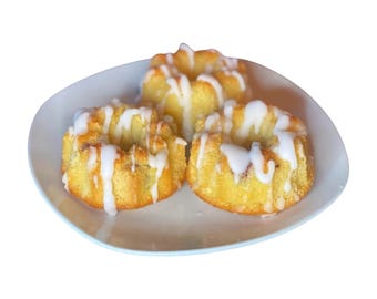 May include: Three mini bundt cakes on a white plate. The cakes are golden brown with a white glaze drizzled over the top. The plate has a slightly irregular shape and a light blue rim. The cakes are arranged in a group.