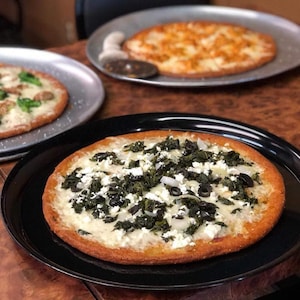May include: Three pizzas on display. The front pizza, on a black plate, has a white sauce base, topped with spinach, black olives, and white cheese. Two other pizzas are on metal trays, one with a similar topping and the other with an orange topping.
