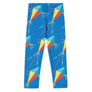 May include: Blue leggings with a repeating pattern of colorful kites. Each kite is drawn with red, yellow, and teal sections, with long, thin tails. The leggings are made of a smooth, stretchy fabric.