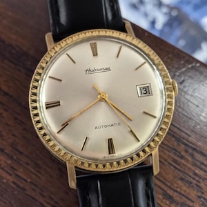 Puede incluir: Un reloj de pulsera automático Habmann dorado con esfera plateada, agujas doradas y ventana de fecha a las 3. El reloj tiene una correa de cuero negro y un bisel texturizado. La esfera indica "AUTOMATIC" e "INCABLOC".