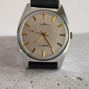 May include: A vintage TEMPIC wristwatch with a silver face and gold hands and markers. The watch has a black leather strap and the text "17 JEWELS" is visible. The watch is set against a grey background.