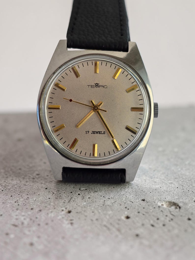May include: A vintage TEMPIC wristwatch with a silver face and gold hands and markers. The watch has a black leather strap and the text "17 JEWELS" is visible. The watch is set against a grey background.