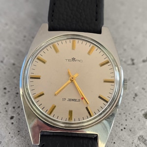 May include: A vintage TEMPIC wristwatch with a silver case and a light-coloured face. The watch features gold hands and markers, with the text "17 JEWELS" and the brand name "TEMPIC". It has a black leather strap.