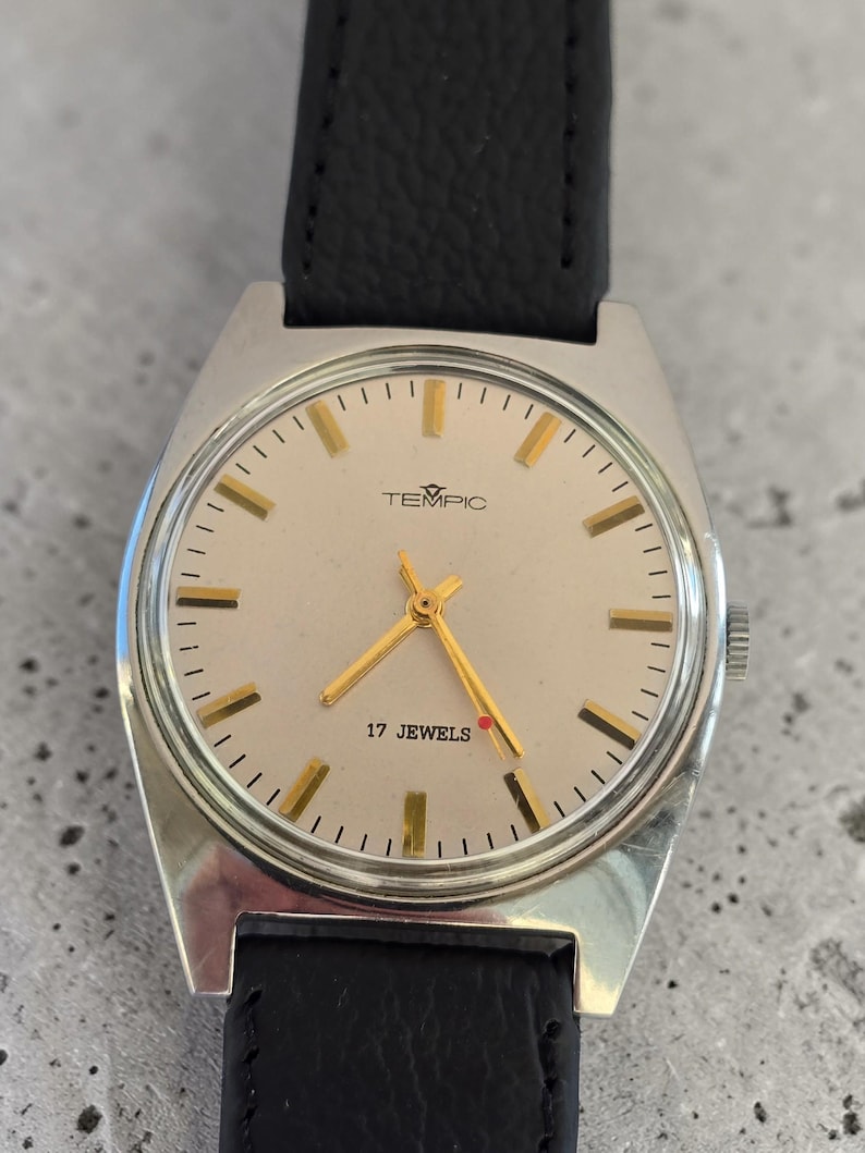 May include: A vintage TEMPIC wristwatch with a silver case and a light-coloured face. The watch features gold hands and markers, with the text "17 JEWELS" and the brand name "TEMPIC". It has a black leather strap.