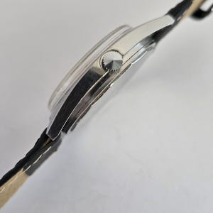 May include: A silver wristwatch with a black leather strap. The watch face is round with a silver bezel and a textured crown. The strap is black with a light beige underside.