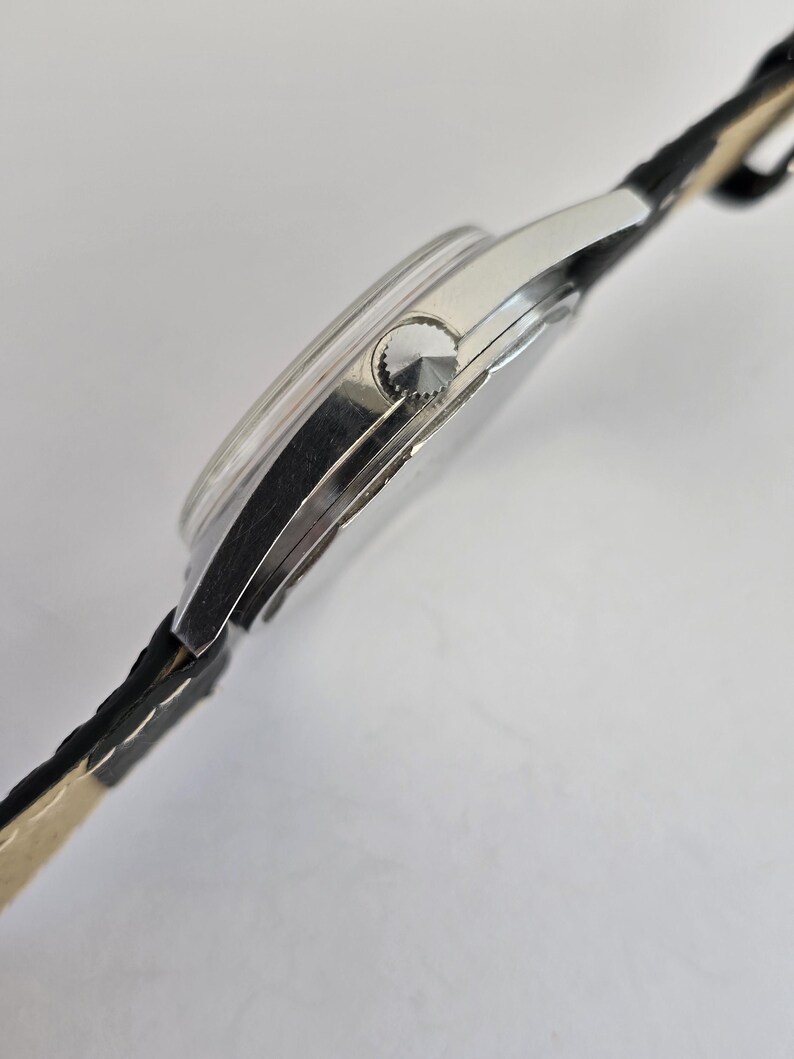 May include: A silver wristwatch with a black leather strap. The watch face is round with a silver bezel and a textured crown. The strap is black with a light beige underside.