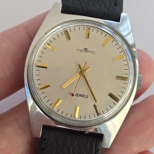May include: Silver TEMPIC wristwatch with a light-coloured face, gold hands and markers, and a black leather strap. The watch face displays the text "TEMPIC" and "17 JEWELS".