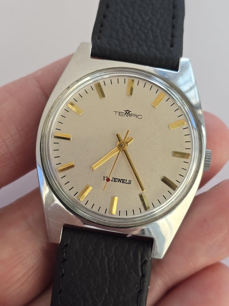 May include: Silver TEMPIC wristwatch with a light-coloured face, gold hands and markers, and a black leather strap. The watch face displays the text "TEMPIC" and "17 JEWELS".