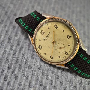 Op de afbeelding: Vintage Jormac 15 Jewels polshorloge met een goudkleurige kast en een crèmekleurige wijzerplaat. Het horloge heeft zwarte cijfers en wijzers en een zwart-groene gestreepte band. Een klassiek horloge.