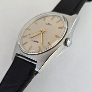 May include: A vintage wristwatch with a silver case and a light-coloured face. The watch has gold hands and markers, with the text "17 JEWELS". It features a black leather strap.