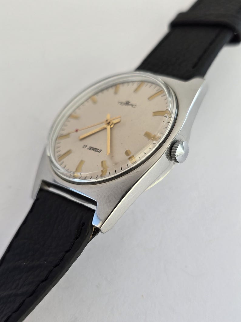 May include: A vintage wristwatch with a silver case and a light-coloured face. The watch has gold hands and markers, with the text "17 JEWELS". It features a black leather strap.