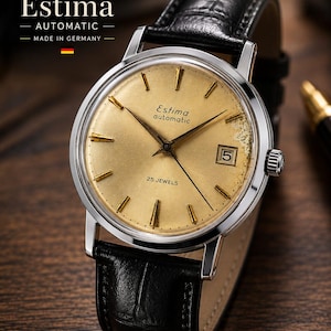May include: An Estima automatic wristwatch with a round silver-tone case and a textured gold-colored dial. The watch features gold-tone hour markers, a date window at the three o'clock position, and a black leather strap with a crocodile pattern.