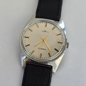May include: A vintage TEMPIC wristwatch with a silver case and a cream face. The watch has gold hands and markers, with the text "TEMPIC" and "1 Jewels". The watch has a black leather strap.